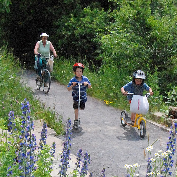 2 young children on scooters and a woman on a bike on a nature trail